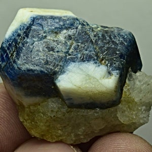 May include: A blue sapphire crystal with a white, opaque, and translucent appearance. The crystal is sitting on a bed of smaller, clear crystals.