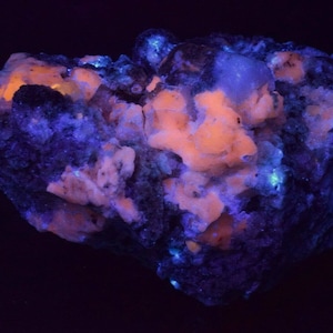 May include: A close-up of a rock with a mix of white, blue, and orange crystals. The rock is illuminated with a UV light, causing the crystals to glow.