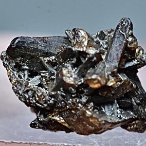 May include: A cluster of dark gray to black, metallic,  crystals with a slightly iridescent sheen. The crystals are sharp and angular, with some showing a slight golden hue.