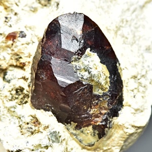 Pode incluir: Um cristal de gema facetado vermelho escuro incrustado em uma matriz rochosa marrom claro.