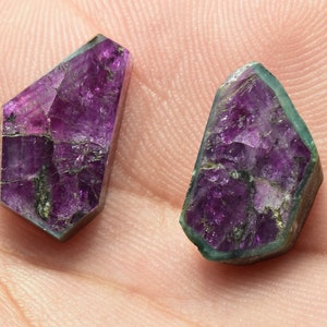 Pode incluir: Duas pedras preciosas roxas e verdes com corte facetado. As pedras preciosas estão sobre uma superfície rosa claro.