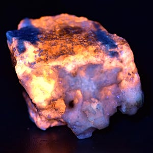 May include: A close-up of a glowing rock with a rough, textured surface. The rock displays a vibrant color palette, with areas of bright orange, yellow, and blue. The overall effect is a striking display of natural mineral beauty.