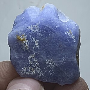 May include: A rough, light blue, opaque gemstone with white and yellow inclusions. The gemstone is irregularly shaped and has a smooth, polished surface.