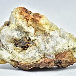 May include: A close-up of a rough, natural mineral specimen. The rock features a mix of colors, including white, tan, brown, and hints of darker shades. The texture appears crystalline and layered, with visible variations in the rock's composition.