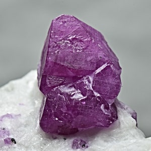May include: A vibrant, deep purple ruby crystal with a faceted, geometric shape. The crystal is resting on a white, textured surface.