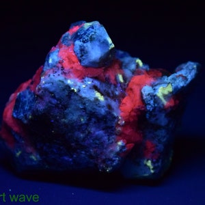 May include: A close-up of a red and yellow fluorescent mineral specimen under short wave UV light. The mineral is a cluster of crystals with a rough, textured surface.