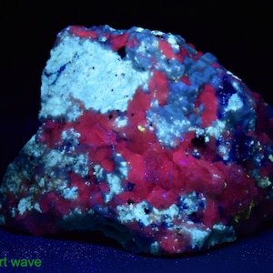 May include: A close-up of a rock sample under short-wave UV light. The rock is a mix of white, pink, and blue minerals. The pink mineral fluoresces brightly under UV light.