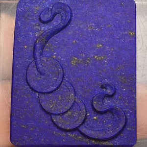 Op de afbeelding: Een diepblauwe lapis lazuli hanger met een gegraveerd ontwerp van een gestileerde slang met een ketting van ronde vormen. De steen heeft vlekken van gouden pyriet.