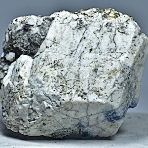 May include: A close-up of a rough, irregularly shaped rock. The rock is primarily white with dark gray and black patches and veins. The surface has a textured appearance with visible cracks and imperfections, suggesting a natural mineral formation.