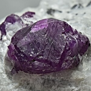 Puede incluir: Un gran cristal de rubí de color púrpura intenso con una superficie rugosa y facetada. El cristal está incrustado en una matriz blanca y cristalina.