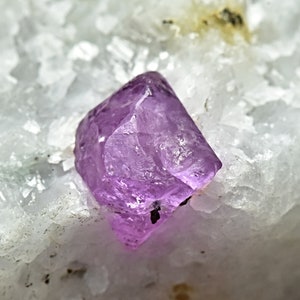 May include: A single, large, purple crystal with a sharp, faceted shape. The crystal is resting on a bed of white, crystalline material.
