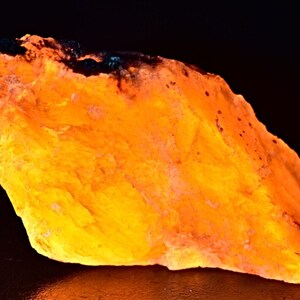 May include: A large, irregularly shaped piece of orange calcite with a dark, almost black, area on one side. The calcite is translucent and glows brightly under UV light.