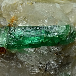 Puede incluir: Un cristal de esmeralda verde vibrante incrustado en una matriz de cuarzo transparente. La esmeralda tiene una forma rectangular distintiva y está rodeada de cristales de cuarzo más pequeños.