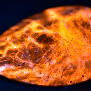 May include: A close-up of a smooth, orange-red stone with a swirling, white pattern. The stone is illuminated with a long-wave UV light, causing it to glow brightly.