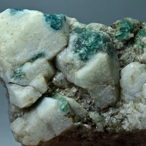 May include: Close-up of a mineral specimen featuring a matrix of white and gray rock with patches of teal-colored crystals. The crystals are clustered within the rock, creating a textured appearance. The mineral has a natural, raw aesthetic.