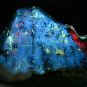 May include: A close-up of a blue, white, and red mineral specimen under UV light. The mineral has a rough, textured surface with a variety of colors and patterns.