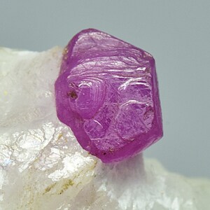 May include: A vibrant pink ruby crystal with a hexagonal shape, set on a white matrix. The crystal has a smooth, polished surface and is highly reflective.