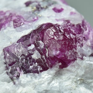 Puede incluir: Primer plano de un cristal de rubí incrustado en una matriz blanca. El rubí es de color rojo intenso y tiene una superficie rugosa y texturizada.