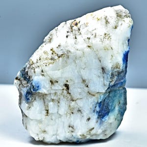 May include: A raw, uncut mineral specimen with a predominantly white base, accented by patches of deep blue and flecks of brown and green. The stone has a rough, textured surface, showcasing its natural, unpolished state. The mineral is likely a type of gemstone.