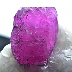 May include: A large, vibrant pink crystal with a rough, faceted surface. The crystal is sitting on a white, textured surface.