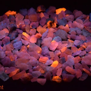 May include: A close-up of a group of small, irregularly shaped, pink, orange, and purple stones. The stones are glowing under a black light, creating a vibrant and colorful display. The stones are arranged in a circular pattern, with the text "Fluorescent" visible at the bottom of the image.