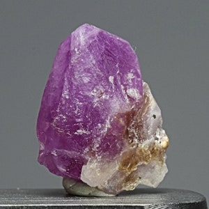 Puede incluir: Un cristal de color púrpura vibrante con una forma puntiaguda y una apariencia translúcida ligeramente nublada. El cristal tiene una superficie rugosa y texturizada y está parcialmente cubierto por un mineral blanco opaco.