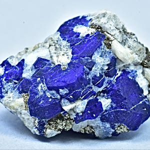 May include: A striking piece of raw lapis lazuli, showcasing deep blue crystalline structures interspersed with white calcite and flecks of pyrite. The stone has a rough, natural texture, with a mix of angular and rounded shapes.