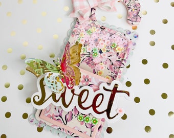 Könnte beinhalten: Ein weißer Türkranz mit einer rosa-weißen Karobinde, einem Blumendesign und einem Schmetterling. Der Kranz hat eine Tasche mit dem Wort "sweet" in goldenen Buchstaben.