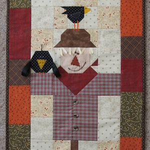 Pode incluir: Uma colcha de retalhos com um espantalho com um chapéu castanho, uma camisa vermelha e calças de xadrez. A colcha é feita de vários tecidos em tons de castanho, vermelho, laranja e branco. Há dois pássaros pretos na colcha, um em cima do chapéu do espantalho e outro à esquerda do espantalho.