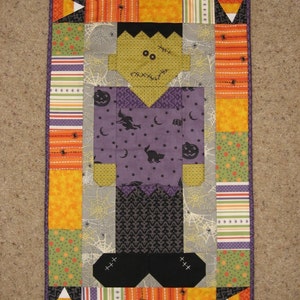 May include: A patchwork quilt block featuring a green, orange, and purple Frankenstein monster with a stitched-on smile. The block is surrounded by a border of orange, green, and white fabric with a candy corn design.