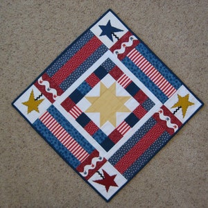 Puede incluir: Una colcha de retazos roja, blanca y azul con un diseño de estrella en el centro. La colcha está hecha de diferentes telas con rayas, lunares y líneas onduladas. La colcha tiene forma cuadrada y un patrón de diamante.