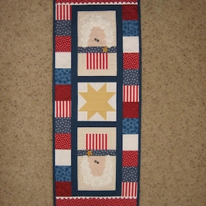 以下が含まれることがあります： 星と2匹の羊のデザインが施された赤、白、青の愛国的なテーブルランナー。羊は赤、白、青のストライプの帽子をかぶっています。