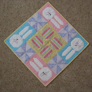 May include: A square Easter-themed quilt featuring four white bunny faces with embroidered details and four decorated Easter eggs. The quilt has a yellow, pink, and blue color scheme with a yellow border.