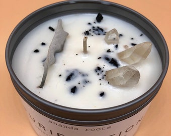 May include: A white candle with a gray lid and the text "ananda roots" and "URIFICATION" with clear quartz crystals, black sage, and black specks embedded in the wax.