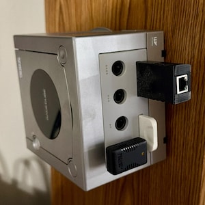 Puede incluir: Consola GameCube plateada con accesorios en blanco y negro adjuntos. La consola tiene una unidad de disco circular y las palabras "GameCube" impresas en la parte delantera. Varios puertos son visibles, con conectores en blanco y negro.