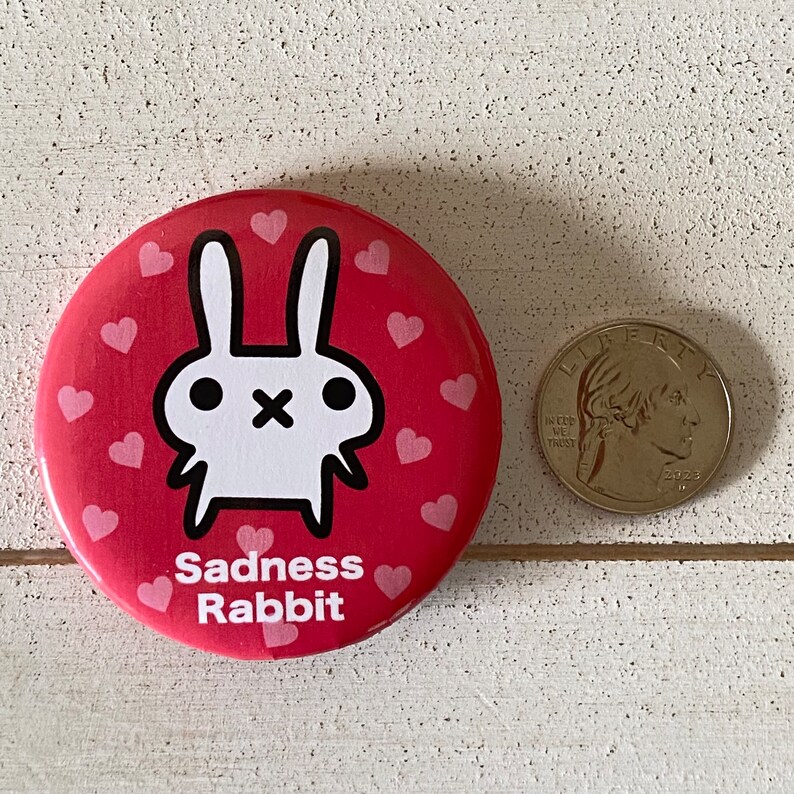 K&ouml;nnte beinhalten: Ein runder, roter Button mit einer wei&szlig;en Kaninchen-Illustration und dem Text "Sadness Rabbit". Der Button ist mit kleinen rosa Herzen verziert. Eine M&uuml;nze liegt neben dem Button zum Gr&ouml;&szlig;envergleich.