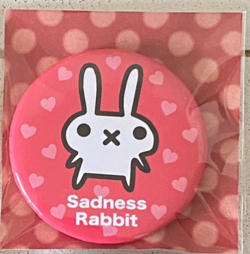K&ouml;nnte beinhalten: Ein runder, roter Button mit einer wei&szlig;en Cartoon-Hasen-Illustration und kleinen rosa Herzen. Der Text "Sadness Rabbit" ist unten auf dem Button in Wei&szlig; aufgedruckt. Der Button ist in einer transparenten Kunststoffverpackung verpackt.