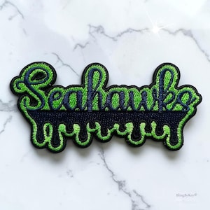 May include: An embroidered patch featuring the word Seahawks in dark blue script with a bright lime green outline. The lower portion of the design consists of a dark blue dripping liquid effect with a bright lime green border and black edges.