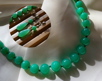 Op de afbeelding: Een ketting met gladde, ronde, levendig groene kralen, vergezeld door een paar bijpassende oorbellen. Elke oorbel bestaat uit een kleine groene kraal bevestigd aan een druppelvormige groene steen met goudkleurige metalen onderdelen.