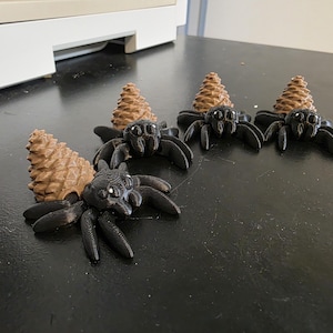May include: Several black spider figurines with brown pinecone-shaped heads are arranged on a dark surface. The spiders have multiple legs and small, detailed eyes. The pinecone tops add a natural element to the design.