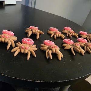 May include: A collection of brown clay spider figurines, each with eight legs and a pink donut on its back. The donuts have white sprinkles. The spiders are arranged on a black surface.