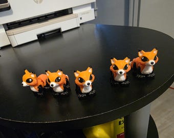 May include: A collection of five orange and white fox figurines with black accents. The foxes are in various poses, all with large, expressive eyes. They are arranged on a black, round table.