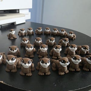 May include: A collection of small, brown otter figurines with white faces and black eyes. The otters are arranged on a dark surface, with some standing and others lying down. The figurines appear to be handmade.