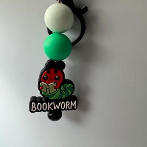 May include: A green, white, and black bookworm keychain with a red bookworm reading a book. The keychain has the word "BOOKWORM" written on it.