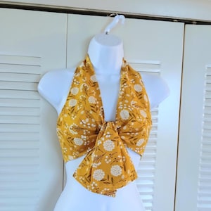 May include: Mustard yellow halter top with a white floral pattern. The top is tied in the front and is displayed on a white mannequin. The fabric has a delicate, feminine design, suitable for warm weather.