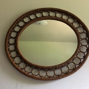May include: A round mirror with a woven brown rattan frame. The frame features a double-layered design with a series of circular openings. The mirror is mounted on a light-colored wall.