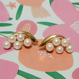 May include: A pair of gold-toned clip-on earrings featuring a curved design. Each earring has five faux pearls. The earrings are set against a floral background with pink and green accents. These earrings are a classic jewelry accessory.
