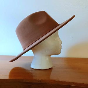 May include: A brown fedora hat with a wide brim, displayed on a white mannequin head. The hat is a solid color and appears to be made of felt or a similar material. The hat is angled, showing the side profile. The background is a light wall and a wooden surface.