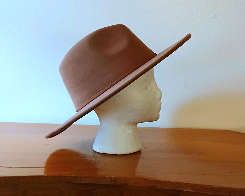 May include: A brown fedora hat with a wide brim, displayed on a white mannequin head. The hat is a solid color and appears to be made of felt or a similar material. The hat is angled, showing the side profile. The background is a light wall and a wooden surface.