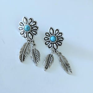 May include: A pair of silver-toned earrings featuring a flower-shaped stud with a central round turquoise stone. Each earring has two dangling silver-toned feather charms attached to the base of the flower, creating a layered, dangling design.